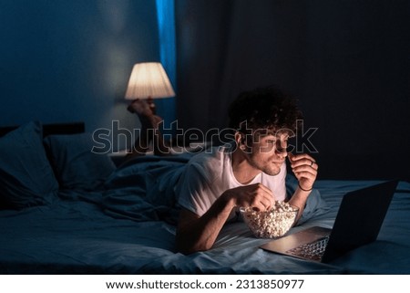 Happy muslim student eating popcorn while lying on bed watching movie on laptop at night at home. Copy space