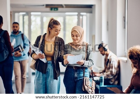 Happy Muslim student and her friend reading their exam results while walking through university hallway.