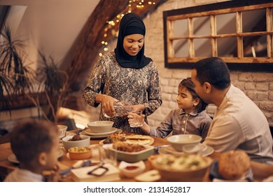 Happy Muslim woman pouring water in daughter's glass while having family dinner at dining table on Ramadan. 