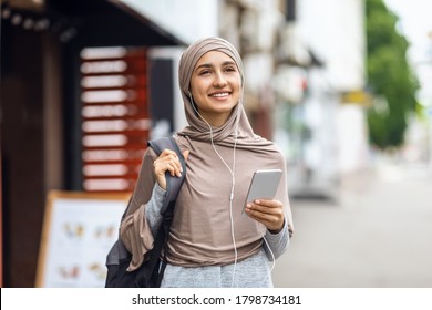 Happy muslim woman walking by street and listening to music, holding smartphone and using earphones, free space - Powered by Shutterstock