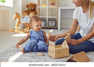 Happy Nanny And Child Playing Game With Wood Blocks Together. Babysitter Looking After Little Kid. Cute Toddler Boy And Mother Sitting On Warm Floor In Modern Nursery Room Building Toy House