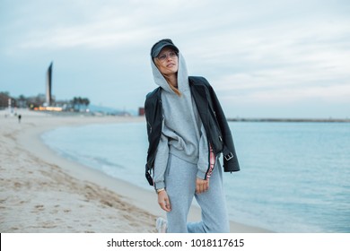 Happy and natural beauty young pretty and attractive woman or teenager wears grey hoodie and sweatpants during workout at beach seaside laughs naturally to camera, during fashion photoshoot influencer
