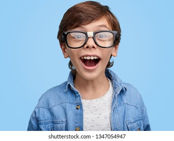 Happy Nerdy Boy In Eyeglasses And Denim Jacket Looking Excitedly At Camera On Blue Background . Closeup Portrait Of Excited School Kid In Broken Glasses