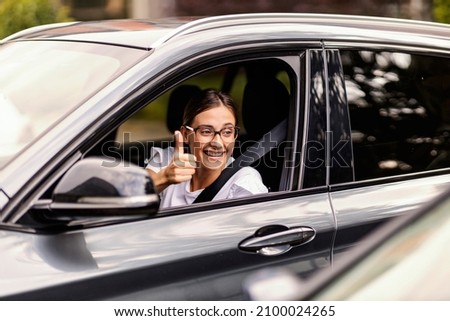 A happy nerdy girl is driving a car and showing thumbs up to other drivers. It is not hard to be polite in traffic. A girl driving a car