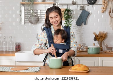 Happy new Asian mom hold and carry baby on baby bag in front and cooking breakfast in the morning. Smile mother carrying little baby infant preparing healthy food.Mom and baby spending time at kitchen - Powered by Shutterstock