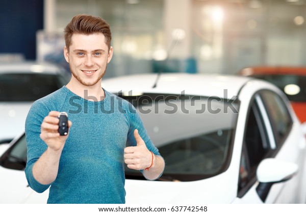 Happy new car owner is showing thumbs\
up and his car key. Car dealership. Focus on the\
face