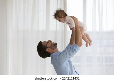 Happy new dad playing with sweet baby, lifting, throwing adorable child up in air, holding kid in arms with love, smiling, laughing, enjoying fatherhood, parenthood. Family concept. Side view