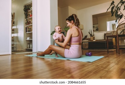 Happy New Mom Working Out With Her Baby At Home. Mother And Baby Smiling Happily While Exercising On An Exercise Mat. Caring Mom Bonding With Her Baby During Her Post-natal Fitness Routine.