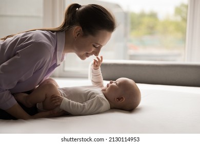 Happy New Mother Caressing, Cuddling Sweet Baby Lying On Back On Bed. Cute Infant Touching Mom Smiling Face. Devoted Mum Playing With Little Child With Love, Affection, Joy, Enjoying Motherhood