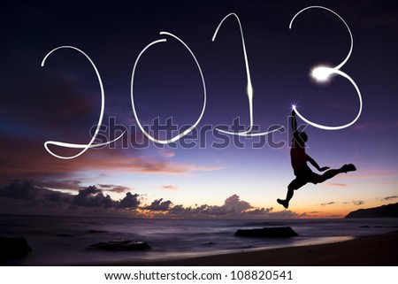 happy new year 2013. young man jumping and drawing 2013 by flashlight in the air on the beach before sunrise