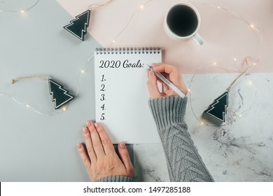 Happy New Year 2020. Woman's hand writing 2020 Goals in notebook decorated with Christmas decorations on the tricolor background. Top view, flat lay