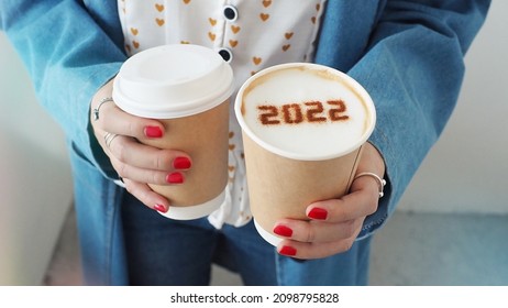 Happy New Year 2022 food art theme female hands holding takeaway disposable double wall paper coffee cups contained cappuccino with number 2022 on frothy surface and another one with plastic lid. - Powered by Shutterstock