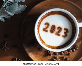 Happy New Year 2023 celebrated coffee cup with number 2023 over frothy surface served on wooden background with coffee beans, star anise, silvery gray foliage of dusty miller plant. Holidays food art. - Shutterstock ID 2232976991