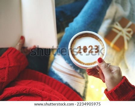 Happy New Year 2023 celebrated coffee cup with number 2023 on frothy surface holding by woman in red knitted sweater with jeans while relaxed sitting on the couch with gift box. Holidays food art.