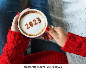 Happy New Year 2023 holidays food art theme, woman in red knitted sweater with jeans holding white coffee cup with number 2023 on frothy surface while relaxed sitting on the couch. (top view) - Shutterstock ID 2217636025