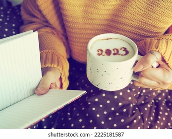 Happy New Year 2023 holidays food art theme, woman in yellow knitted sweater with brown and white polka dot blanket holding white coffee cup with number 2023 on frothy surface while reading a book. - Powered by Shutterstock