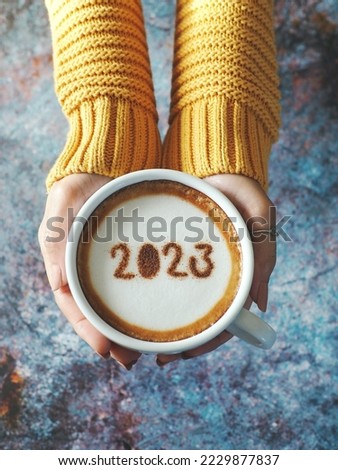 Happy New Year 2023 theme number 2023 on frothy surface of cappuccino served in white coffee mug holding by female hands over rustic blue background. Holidays food art, new year new you. (top view)