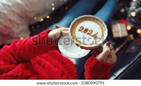 Happy New Year 2024 celebrated coffee cup with number 2024 on frothy surface holding by woman in red knitted sweater with jeans while relaxed sitting on the couch with gift box. Holidays food art.