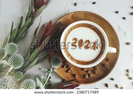 Happy New Year 2024 theme coffee cup with number 2024 over frothy surface served on wooden saucer and white cement background with coffee beans, Safari Sunset Dark Red, green plant. Holidays food art.