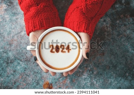 Happy New Year 2024 theme number 2024 on frothy surface of cappuccino served in white coffee mug holding by female hands over rustic blue background. Holidays food art, new year new you. (top view)