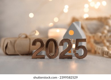 Happy new year 2025 postcard. Golden digits in front of Christmas lights, candles, gift box. White wooden background. Copy space for greeting text
