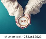 Happy New Year 2025 theme number 2025 over frothy surface of cappuccino served in white coffee mug holding by female hands on green background. Holidays food art, new year new you. (top view)