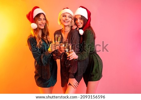 Happy New Year. Three happy girls in dresses and Santa Claus hats holds glasses of champagne having fun on colorful background. Merry Christmas. New Year party. 