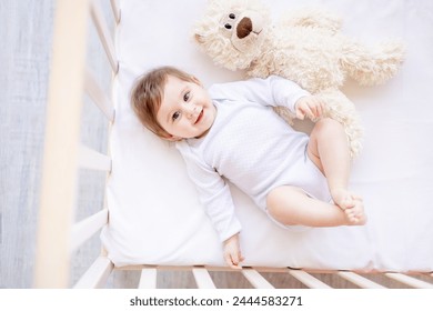Una niña recién nacida feliz en una cuna blanca en un dormitorio brillante en primer plano con ropa blanca con un oso de peluche suave, sonriendo o riendo, un lugar para el texto, vista superior