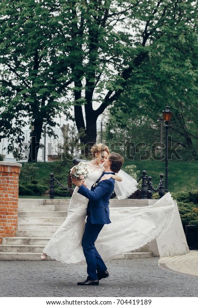 Happy newlywed couple portrait, strong\
handsome groom lifting up bride in the air, groom carrying\
beautiful bride outdoors, car in the\
background