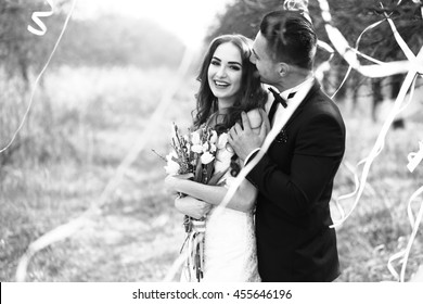 Happy Newlywed Couple Posing And Smiling In Wedding Dress And Suit With Flowers In Forest Decorated With Ribbons Outdoor, Black And White