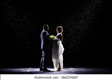 Happy Newlywed Couple Standing Against The Background Of Falling Snow. A Man In A Suit And A Woman In A White Long Dress With A Bouquet On A Black Background In The Rays Of Studio Light.