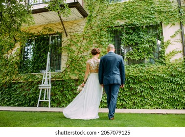 Happy newlyweds hold hands and walk on lawn against background of entwined by lianas house. Back view.