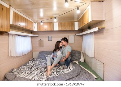 Happy Newlyweds Hug And Kiss Inside The Trailer Track On The Couch. A Honeymoon Trip Across The Country. Budget Vacation For A Young Family. Beautiful Couple Relaxing In The Camping. Concept Caravan