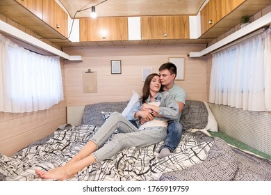 Happy Newlyweds Hug And Kiss Inside The Trailer Track On The Couch. A Honeymoon Trip Across The Country. Budget Vacation For A Young Family. Beautiful Couple Relaxing In The Camping. Concept Caravan