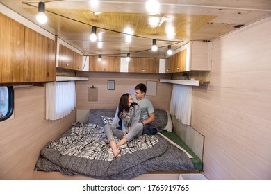 Happy Newlyweds Hug And Kiss Inside The Trailer Track On The Couch. A Honeymoon Trip Across The Country. Budget Vacation For A Young Family. Beautiful Couple Relaxing In The Camping. Concept Caravan