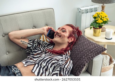 Happy non-binary person with pink hair making a phone call, lying on a comfortable sofa at home, enjoying a relaxing moment with a body positive attitude and celebrating their identity