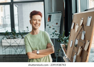 happy non-binary person with short hair looking at camera near corkboard with paper notes in office