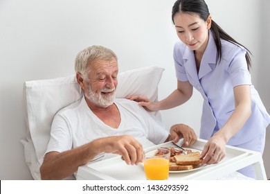 Happy Nurse Assist And Help Elderly Senior Man To Eat Breakfast On Bed In Bedroom At Nursing Home