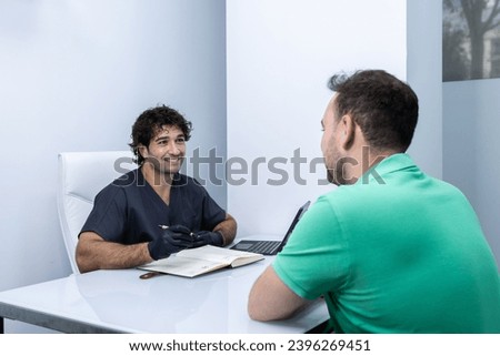 A happy nurse sits across from his client as they talk about the treatment to follow. He has got his diary and the digital tablet on the table office.