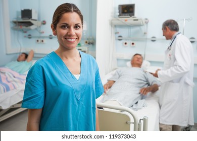 Happy Nurse Standing In Hospital Room With Doctor And Patient Talking In Background, Healthcare Workers In The Coronavirus Covid19 Pandemic