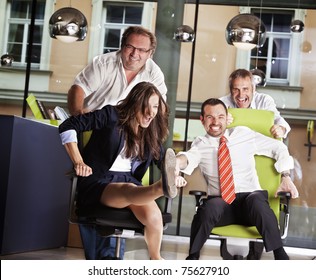 Happy Office Employees Having Fun At Work In An Office Chair Race.