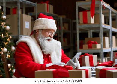 Happy Old Bearded Santa Claus Wearing Costume Holding Cell Phone Using Mobile Christmas Promotion Sale App On Smartphone Video Calling In Virtual Online Chat Sitting At Table In Workshop On Xmas Eve.