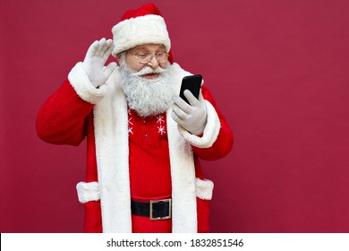 Happy Old Bearded Santa Claus Wearing Costume Waving Hand Holding Cell Phone Using Mobile App Video Calling Greeting On Merry Christmas On Smartphone In Virtual Online Chat Isolated On Red Background.