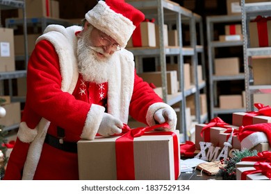 Happy Old Bearded Santa Claus, Saint Nicholas Packing Present Box Wrapping Gift With Red Ribbon Preparing Post Shipping Delivery Parcel On Table In Workshop. Merry Christmas Shipping Delivery Concept.