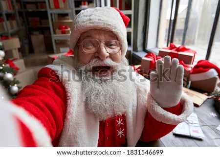 Happy old bearded Santa Claus wearing costume holding phone waving hand taking selfie, video calling, recording video Merry Christmas greeting or shooting vlog standing in workshop, face camera view.
