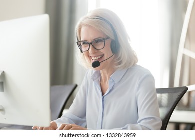 Happy old businesswoman in headset speaking by conference call looking at computer, mature female aged call center agent operator telemarketer talking consulting customer service support in office