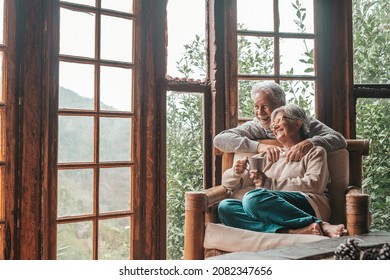 Happy old caucasian couple spending leisure time looking out through window at home. Loving husband embracing wife from behind sitting on armchair at house and admiring view from transparent door  - Powered by Shutterstock