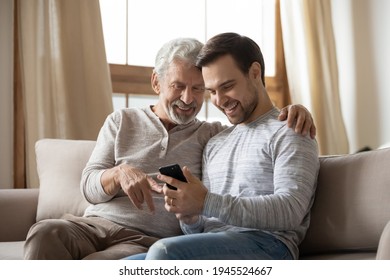 Happy Old Caucasian Father And Adult Grownup Son Sit On Sofa At Home Use Modern Smartphone Gadget Together. Smiling Senior Dad And Millennial Man Kid Look At Cellphone Screen Talk On Video Call.