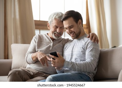 Happy old Caucasian father and adult grownup son sit on sofa at home use modern smartphone gadget together. Smiling senior dad and millennial man kid look at cellphone screen talk on video call. - Powered by Shutterstock