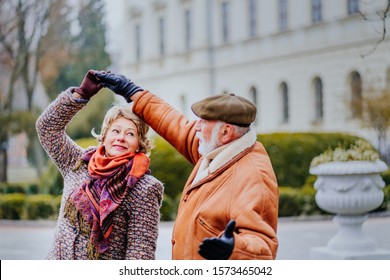 Happy Old Couple Dancing At Autumn Park. Senior Ma Flirting With Blonde Elderly Woman Outdoors In City Square In Cold Weather City Street. My Love Let's Dance Together. Lifestyle Invitation Concept.